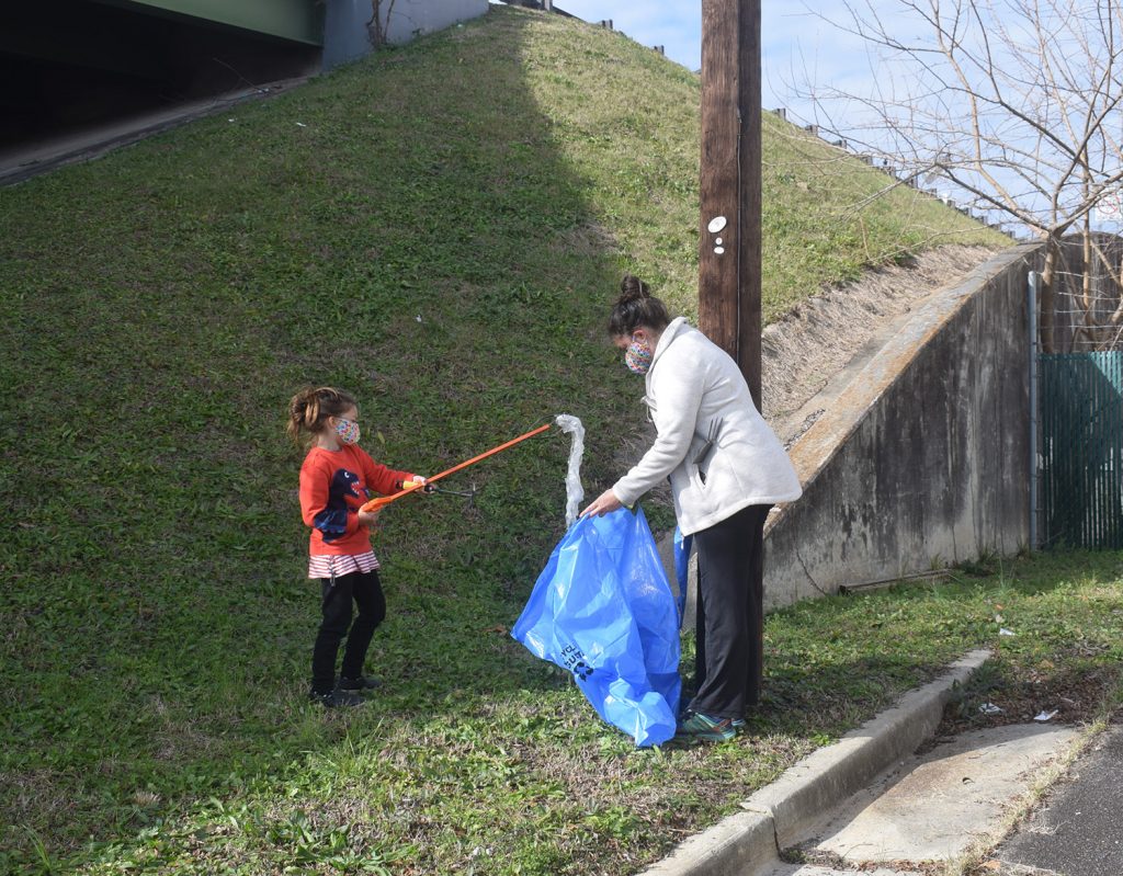 Volunteers pick up trash under the John C. Calhoun Expressway. Pictured is a child with a trash stick, putting debris into a bag that his guardian holds.